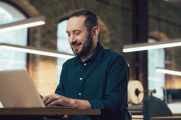 Wesoły, entuzjastyczny pracownik siedzący przy stole w wydawnictwie i uśmiechający się podczas korzystania z nowoczesnego laptopa