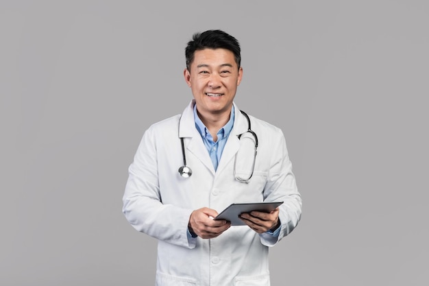Wesoły lekarz azjatycki mężczyzna w średnim wieku w białym fartuchu z tabletem stetoskopowym na białym tle na szarym tle