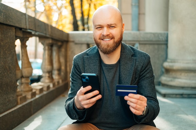 Wesoły łysy mężczyzna korzysta z bankowości mobilnej, trzymając telefon i kartę