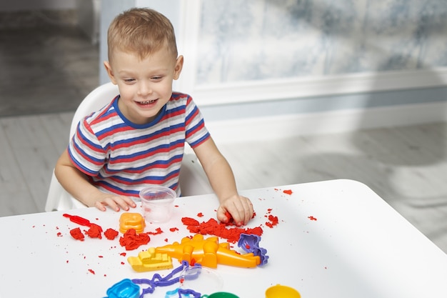 Wesoły mały chłopiec w jasnej koszulce bawi się piaskiem kinetycznym w edukacyjną grę dla dzieci