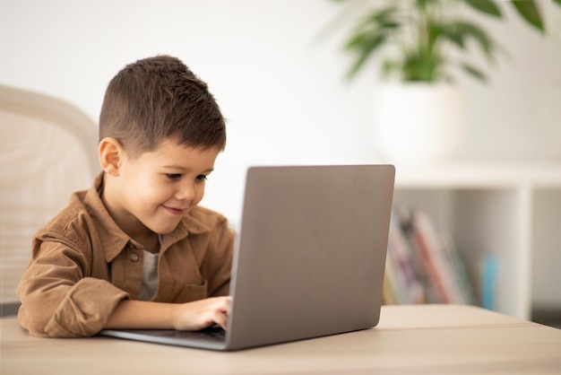 Wesoły mały europejski chłopiec siedzi przy stole, pisząc na komputerze, oglądaj wideo we wnętrzu pokoju