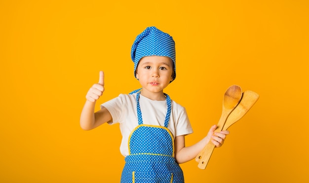 Wesoły mały kucharz w kapeluszu i fartuchu trzyma drewnianą łopatkę i łyżkę na żółtej powierzchni z miejscem na tekst