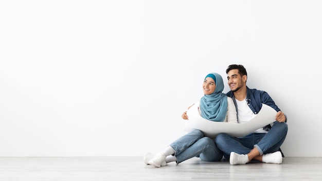 Wesoły mąż i żona z Bliskiego Wschodu w hidżabie, siedzący na podłodze w pustym pokoju, trzymający schemat mieszkania i patrzący na miejsce na reklamę, białe tło ściany, panorama