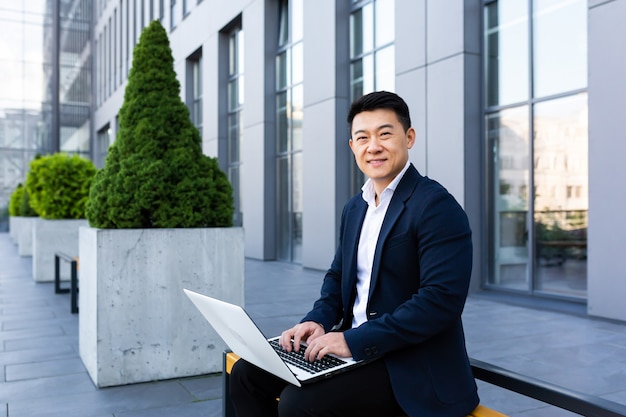 Wesoły mężczyzna azjatycki biznesmen pracujący na laptopie bankowości internetowej na komputerze w pobliżu centrum biurowego, patrząc na kamerę i uśmiechając się