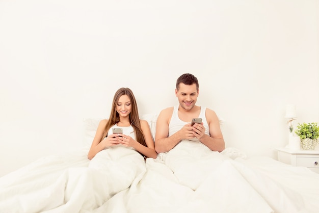 Wesoły mężczyzna i kobieta, leżąc w łóżku z telefonami i rozmawiając