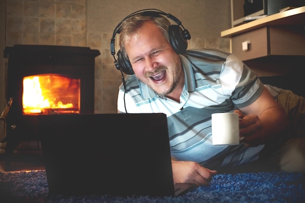 Wesoły mężczyzna ogląda w nocy treści online na laptopie na podłodze pokoju przy kominku