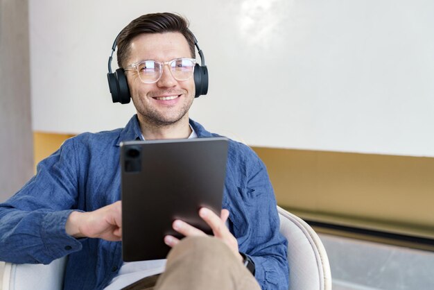 Wesoły mężczyzna z słuchawkami używa tabletu siedzącego wygodnie emitującego zrelaksowany