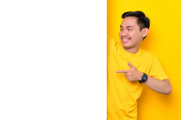 Wesoły młody Azjata w casualowej koszulce, wskazując na białą tablicę reklamową na żółtym tle Koncepcja billboardu promocyjnego