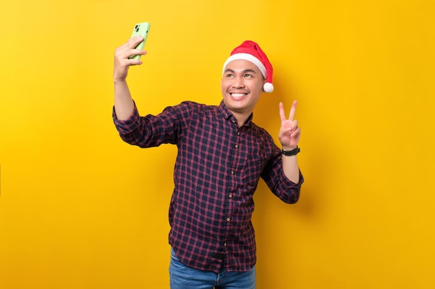 Wesoły młody Azjata w czapce Mikołaja robi selfie na smartfonie pokazując znak zwycięstwa na żółtym tle studia Szczęśliwego Nowego Roku 2023 celebracja wesołych świąt koncepcja
