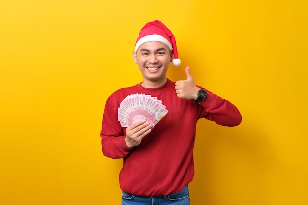 Wesoły młody Azjata w kapeluszu Mikołaja trzymający fana gotówki pokazujący gest kciuka w górę na żółtym tle studia obchody świąt Bożego Narodzenia i koncepcja Nowego Roku