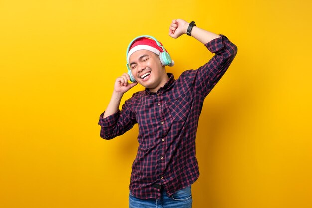 Wesoły młody Azjata w kapeluszu Świętego Mikołaja słuchający muzyki ze słuchawkami i tańczący na żółtym tle studyjnym Szczęśliwego Nowego Roku 2023 koncepcja wesołych świąt