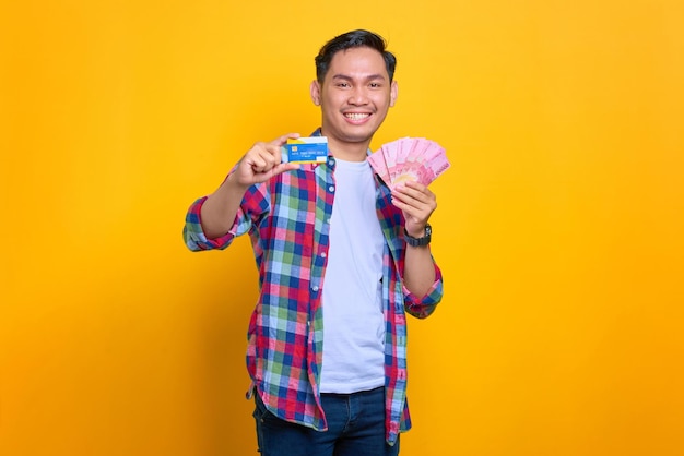Wesoły młody Azjata w koszuli w kratę pokazujący banknoty pieniędzy i kartę kredytową na białym tle na żółtym tle