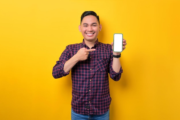 Wesoły młody Azjata wskazujący palcem wskazującym na pusty ekran smartfona odizolowany na żółtym tle Reklama nowej koncepcji makiety aplikacji mobilnej