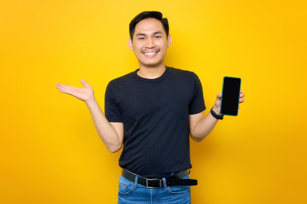 Wesoły młody azjatycki człowiek w swobodnym tshirt pokazując telefon komórkowy z pustym ekranem pokazującym miejsce na kopię na dłoni, polecając aplikację mobilną na białym tle na żółtym tle