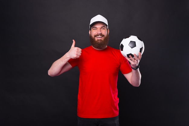 Wesoły młody brodaty mężczyzna pokazuje kciuk gest i trzyma piłkę nożną
