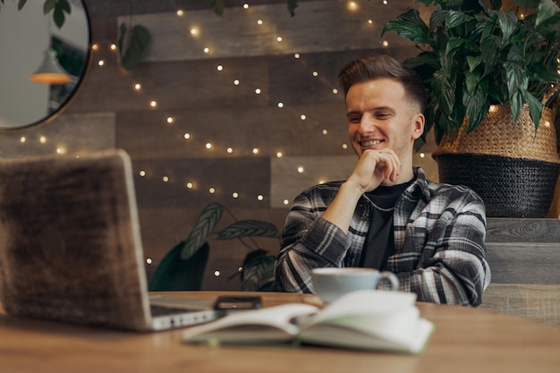 Wesoły młody mężczyzna freelancer w hipsterskiej odzieży pracujący zdalnie na laptopie nad projektem w kawiarni