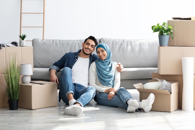 Wesoły młody muzułmański mąż i żona w hidżabie pozują podczas przeprowadzki do nowego mieszkania, siedząc na podłodze przy kanapie wśród papierowych pudełek z rzeczami, obejmując i uśmiechając się do kamery, kopiując przestrzeń