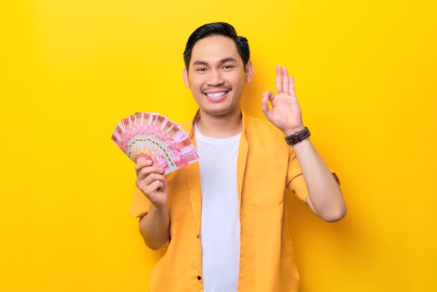 Wesoły młody przystojny Azjata trzymający garść pieniędzy i pokazujący znak porządku na białym tle na żółtym tle