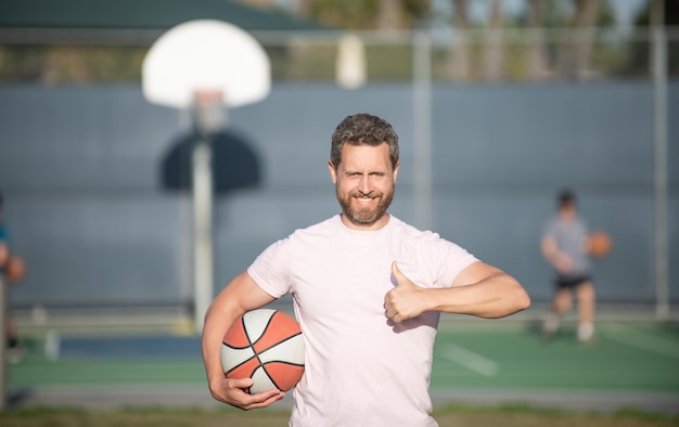 Wesoły muskularny mężczyzna lekkoatletka koszykarz trzymać piłkę odkryty wyświetlone kciuk gest, sukces sportu.