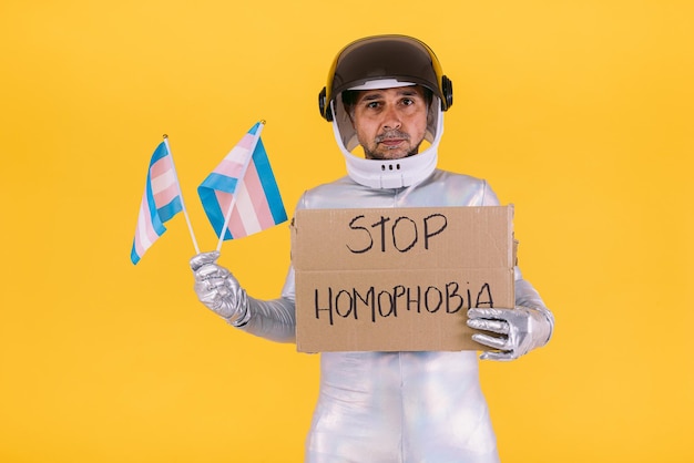 wesoły przebrany za astronautę w hełmie i srebrnym garniturze trzymający dwie małe flagi kolektywu trans i napis Stop homofobii na żółtym tle