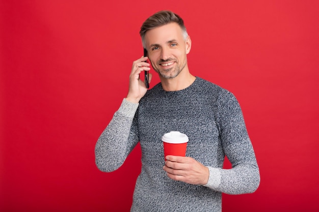 Wesoły siwy facet w swetrze pić kawę z smartphone na czerwonym tle, komunikacja.