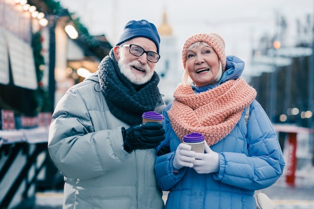 Wesoły starszy mężczyzna i kobieta w ciepłych zimowych ubraniach, stojąc z filiżankami kawy na wynos i patrząc z uśmiechem. Jarmark bożonarodzeniowy w tle