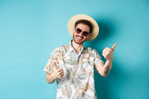 Wesoły turysta bawiący się na wakacjach, pokazujący kciuki do góry i uśmiechnięty, stojący w hawajskiej koszuli i okularach przeciwsłonecznych na niebieskim tle.