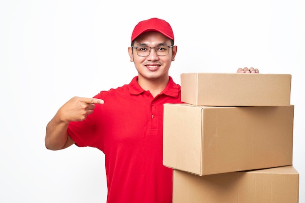 Wesoły uśmiechnięty dostawca w czerwonej koszuli i czapce stojący i wskazujący stosy pudełek z paczkami