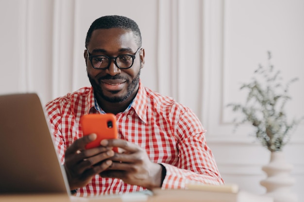 Wesoły uśmiechnięty pracownik biurowy afrykańskiego pochodzenia etnicznego siedzący przy biurku z otwartym laptopem, trzymający smartfona na czacie z przyjaciółmi podczas zdalnej pracy online z domu Koncepcja Freelance