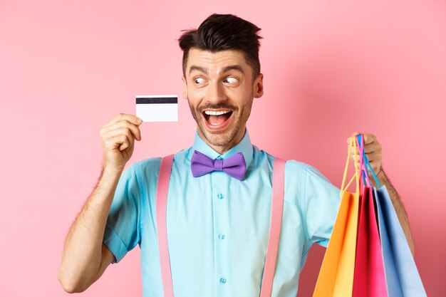 Wesoły zabawny facet patrząc na plastikową kartę kredytową i uśmiechając się, kupując prezenty, trzymając torby na zakupy, stojąc na różowo.