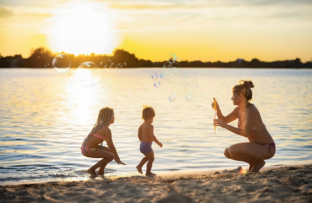 Wesoły Zabawny Szczęśliwy Młodszy Brat I Starsza Siostra Bawią Się Ze Swoją Ukochaną Piękną Wesołą Mamą W Stroju Kąpielowym Bikini, Dmuchając W Jasne Wielokolorowe Bańki Mydlane Na Brzegu Nieśmiałego Jeziora