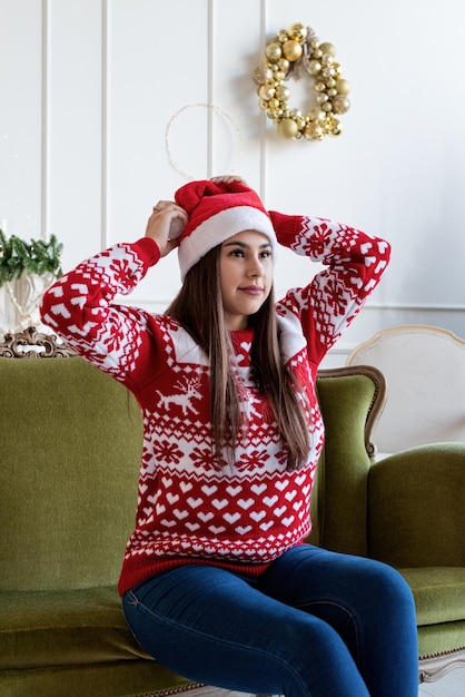 Wesołych Świąt i Szczęśliwego Nowego Roku. Młoda brunetka w santa hat siedzi samotnie na zielonej kanapie w udekorowanym na Boże Narodzenie salonie