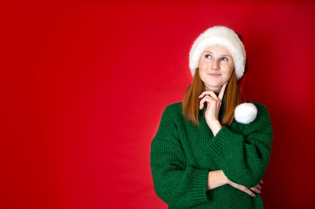 Wesołych Świąt Portret pięknej marzycielskiej nastolatki w przytulnym zielonym swetrze z dzianiny i czapce Mikołaja Czerwone tło to miejsce na tekst