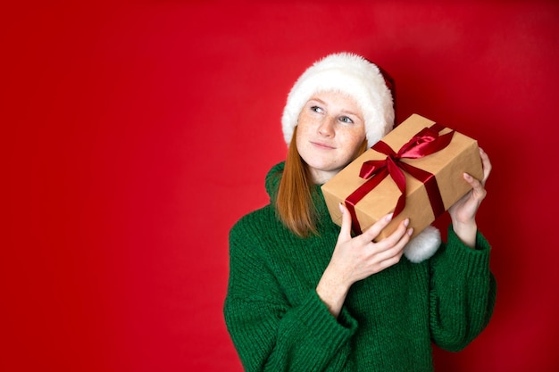 Wesołych Świąt Portret pięknej młodej nastolatki w przytulnym zielonym swetrze z dzianiny i kapeluszu Świętego Mikołaja trzymającym pudełka na prezenty Czerwone tło to miejsce na tekst