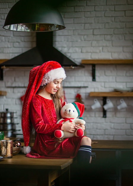 Wesołych Świąt. Śliczna mała dziewczynka w Santa kapeluszu trzyma misia siedzi w kuchni i czeka wakacje.