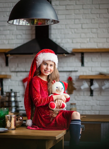 Wesołych Świąt. Śliczna mała dziewczynka w Santa kapeluszu trzyma misia siedzi w kuchni i czeka wakacje.