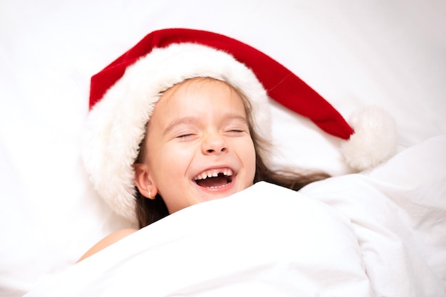Wesołych Świąt. Śmieszna dziewczyna w czapce Mikołaja wygląda spod kołdry w łóżku. Biały