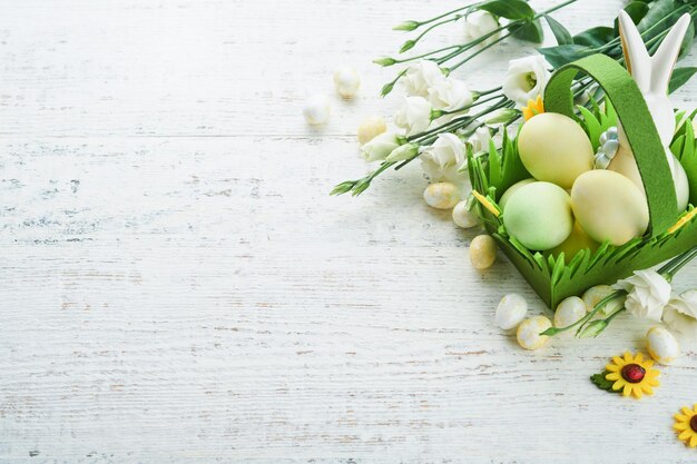 Wesołych Świąt Wielkanocnych jajka i królik w zielonym koszu na białym starym drewnianym stole z białymi i żółtymi różami Wiosna Wesołych Świąt Kartka świąteczna Tło wielkanocne z miejsca na kopię Widok z góry