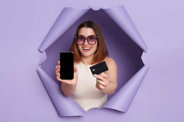 Wewnątrz ujęcie kobiety przebijającej się przez fioletową papierową ścianę i pokazującej kartę kredytową i telefon komórkowy z pustym wyświetlaczem na reklamę ubraną w białą koszulę i okulary przeciwsłoneczne