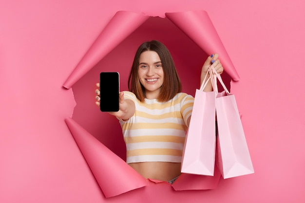 Wewnątrz ujęcie podekscytowanej niezwykle szczęśliwej młodej dziewczyny w pasiastej koszulce przedzierającej się przez papierową dziurę na różowym tle, pokazującej telefon komórkowy z pustym wyświetlaczem i trzymającą torby na zakupy