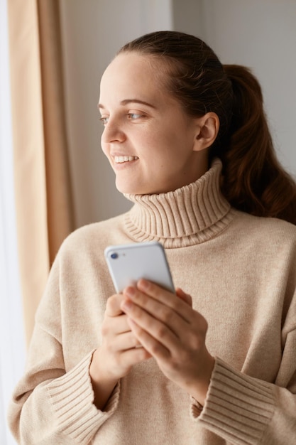 Wewnątrz ujęcie zadowolonej młodej, dorosłej, ciemnowłosej kobiety rasy kaukaskiej z kucykiem, ubranej w beżowy sweter, trzymającej inteligentny telefon i oddalającej się z tajemniczym uśmiechem