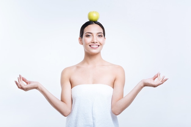 Wewnętrzny spokój. Piękna wesoła młoda kobieta w ręczniku medytuje i trzyma jabłko na głowie, pozując przy białej ścianie