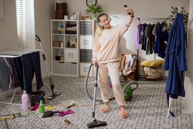 Zdjęcie wideorozmowa podczas sprzątania w domu kobieta trzyma telefon w dłoni i uśmiecha się do kamery robi sobie selfie rozmawia z koleżanką słucha muzyki na dużych słuchawkach bezprzewodowych odkurza mieszkanie