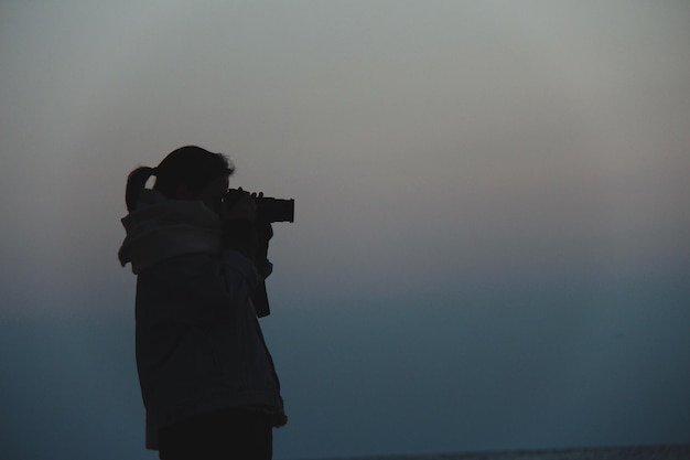 Zdjęcie widok boczny kobiety fotografującej kamerą na tle nieba podczas mglistego zachodu słońca