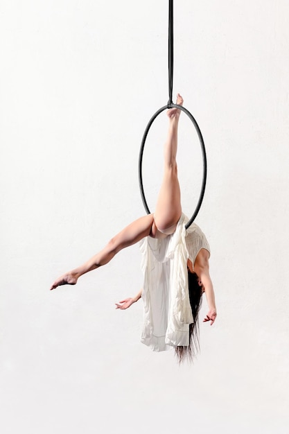 Widok całego ciała z boku elastycznej, szczupłej, bosej akrobatycznej kobiety ćwiczącej na obręczy powietrznej na białym tle w jasnym studio