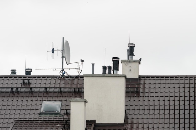 Widok dachu budynku z zamontowanymi antenami i rurami wentylacyjnymi na tle szarego nieba