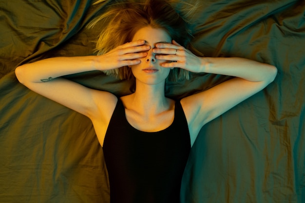 Widok młodej blond zrelaksowanej kobiety w czarnym trykocie zasłaniającym oczy rękami leżąc na łóżku rano niechętnie się obudzić