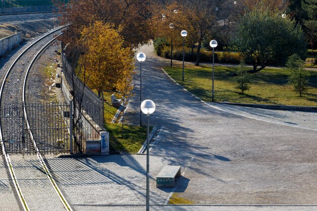 Zdjęcie widok na jesienny park przedstawiający drogę z latarniami oświetlonymi blaskiem świtu po lewej stronie tory kolejowe rysują zakręt niemal równolegle do drogi