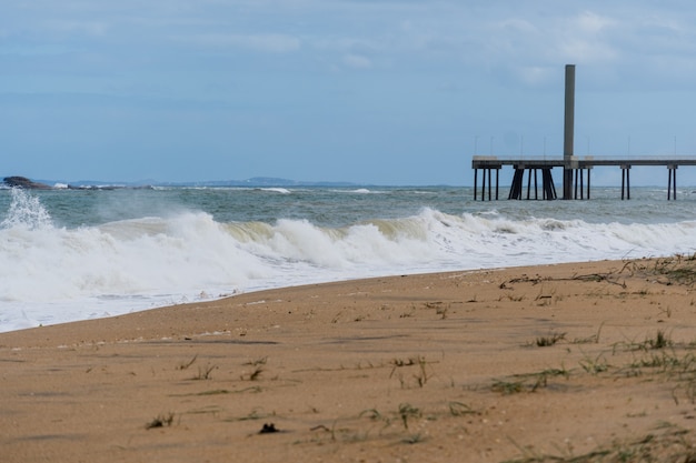 Widok na plażę Rio das Ostras w Rio de Janeiro z molo emisariusza. Słoneczny dzień, błękitne niebo i trochę chmur. Silne morze, żółtawy piasek i mnóstwo skał.