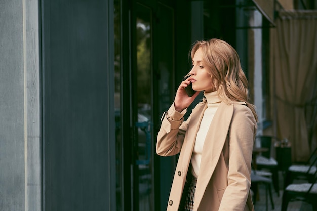 Widok z boku atrakcyjnej blond dziewczyny w stylowym trenczu w zamyśleniu rozmawiającej na telefonie komórkowym na świeżym powietrzu